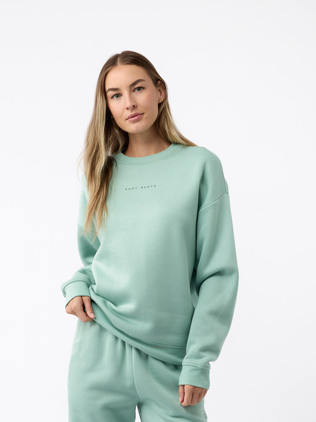 A person with long hair stands in a relaxed posture against a plain white background, wearing the Women's CityScape Crewneck from Cozy Earth in a matching teal set. The crewneck features text on it. 