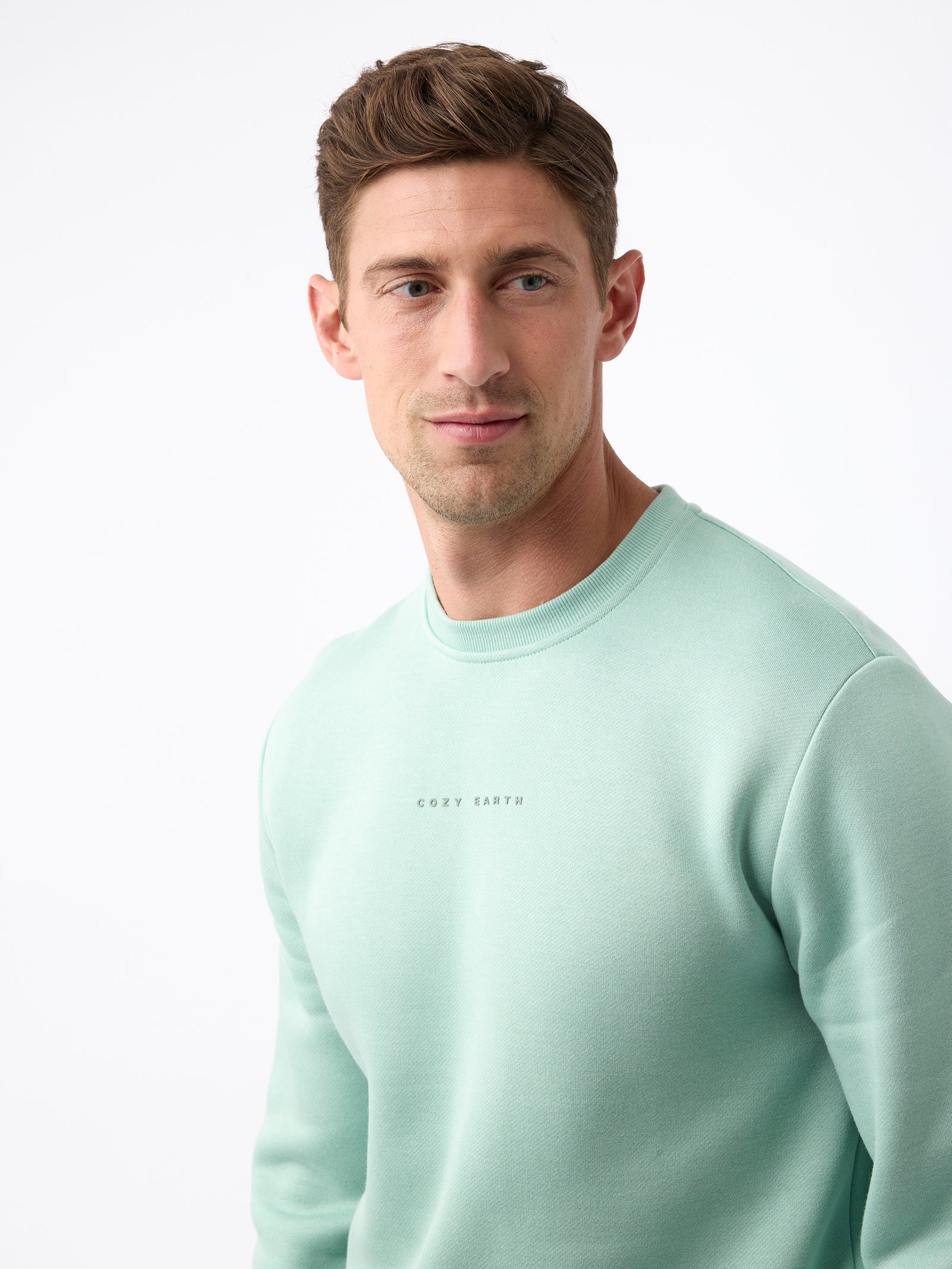 A man with short brown hair is wearing a mint green Men's CityScape Crewneck from Cozy Earth, featuring the brand name printed on the front. He gazes slightly to the side against a plain white background. 