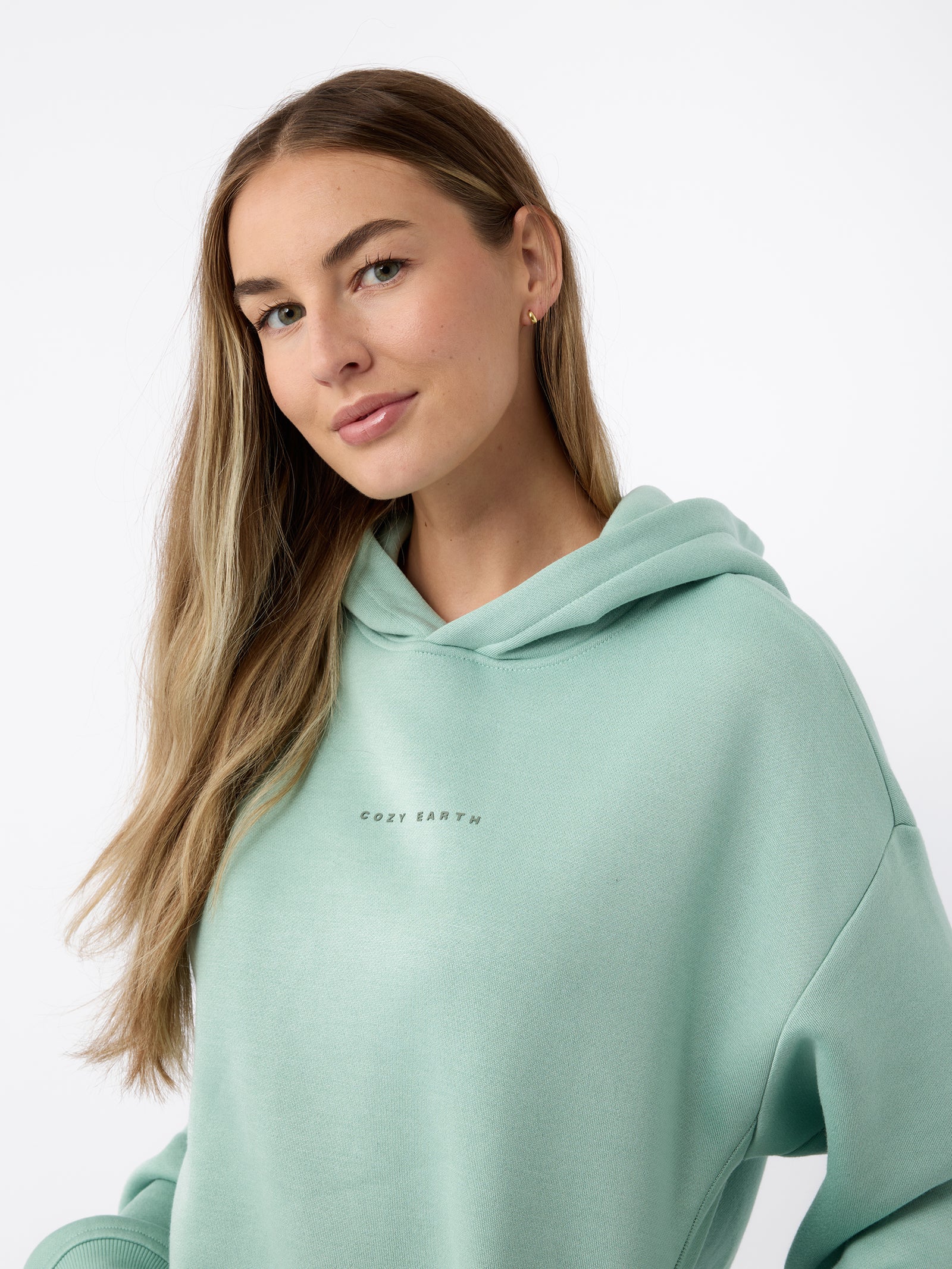 A person with long, light brown hair is wearing a mint green Women's CityScape Hoodie by Cozy Earth. They are smiling softly and standing against a plain white background. 