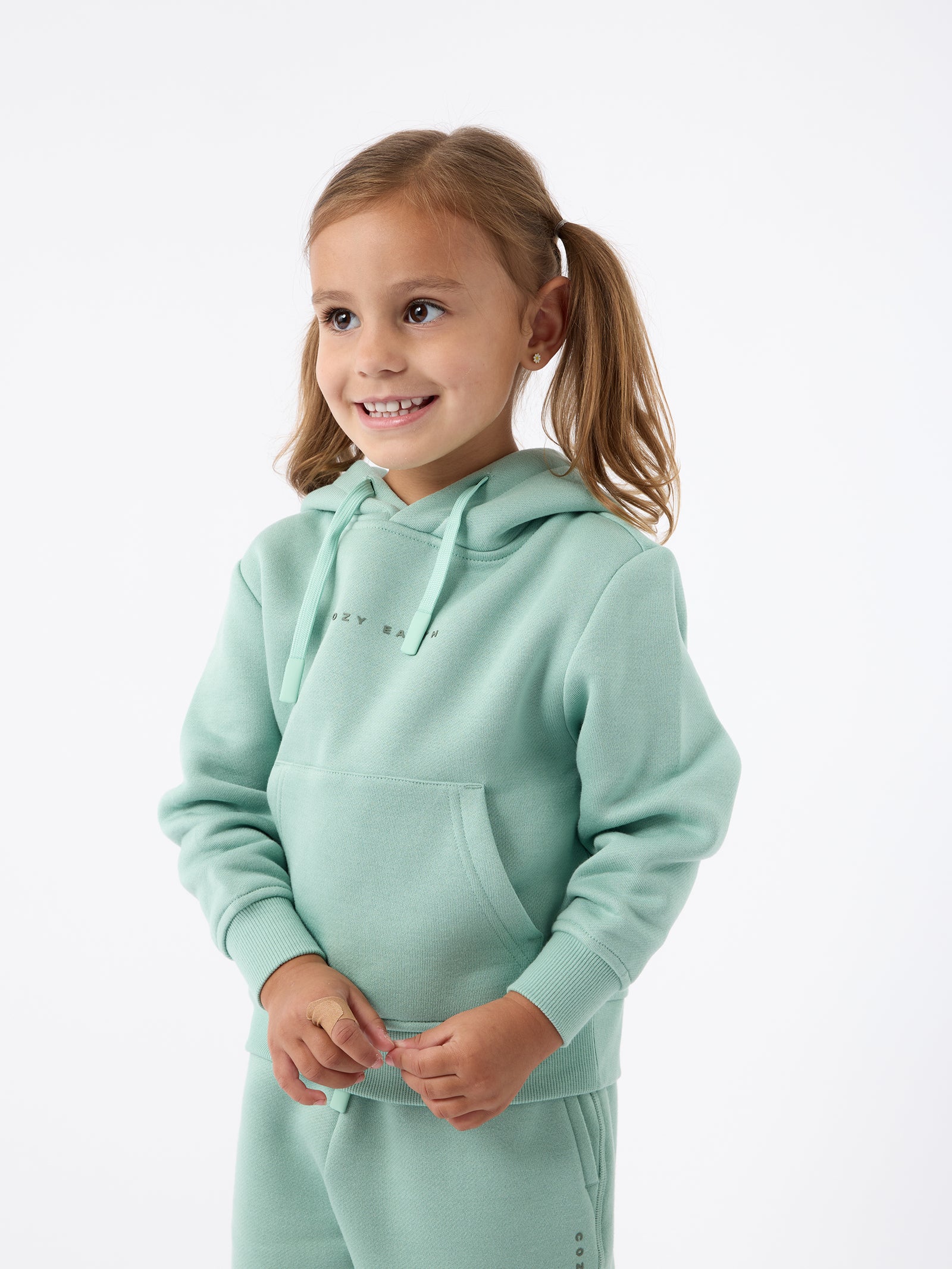 A young girl with light brown hair in pigtails smiles while wearing a mint green Kid's CityScape Hoodie and matching pants from Cozy Earth. She stands against a plain white background. 