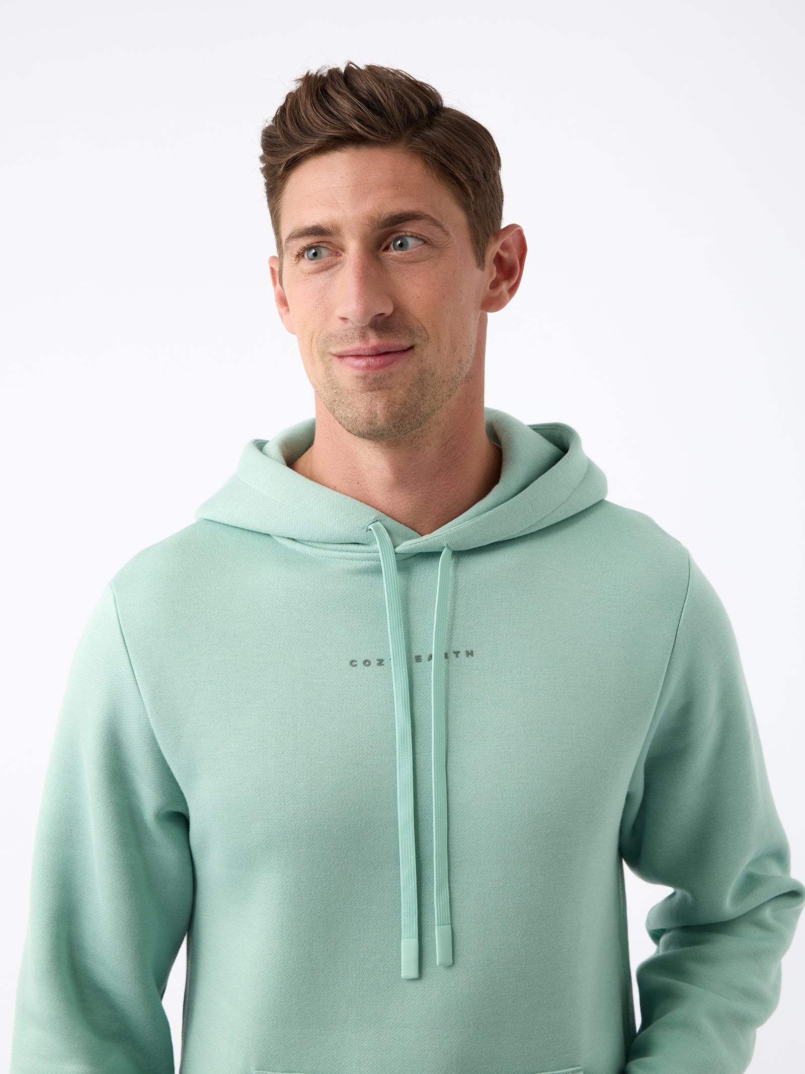 A person wearing the Men's CityScape Hoodie by Cozy Earth stands against a plain white background. The light green hoodie features drawstrings and subtle text on the chest. The person, with short brown hair, is looking slightly to the side while smiling gently. 