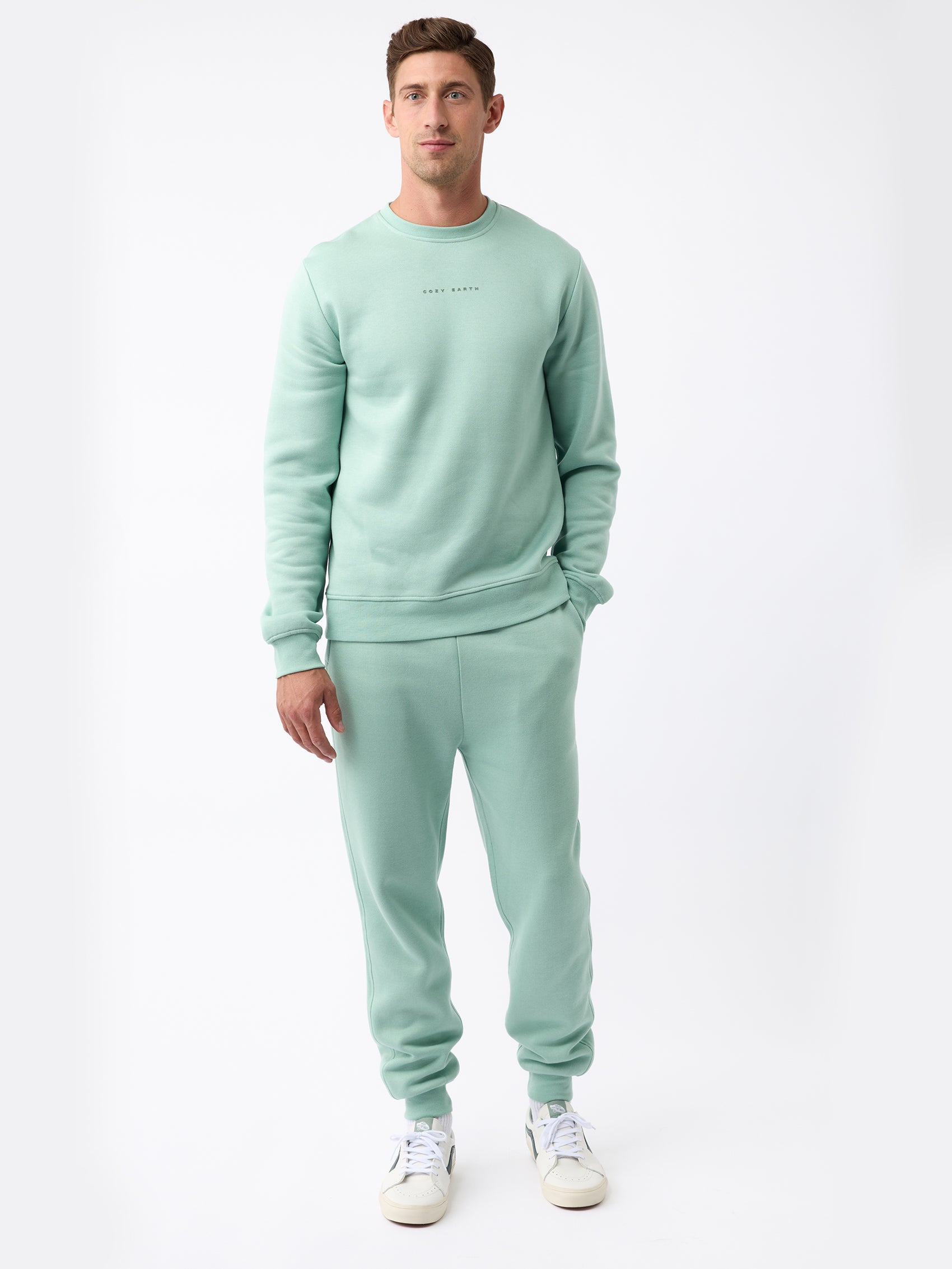 A man is standing against a plain white background, wearing the Men's CityScape Jogger set from Cozy Earth in light green, paired with white sneakers. His hands are relaxed by his sides, and he has short brown hair. |Color:Wintergreen