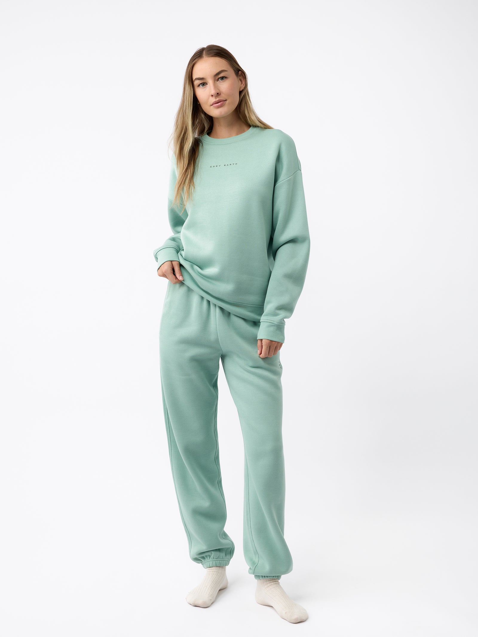 A person in a relaxed pose is wearing the Women's CityScape Sweatpant from Cozy Earth in a matching mint green set, complemented by their long hair and white socks, all against a plain white background. 