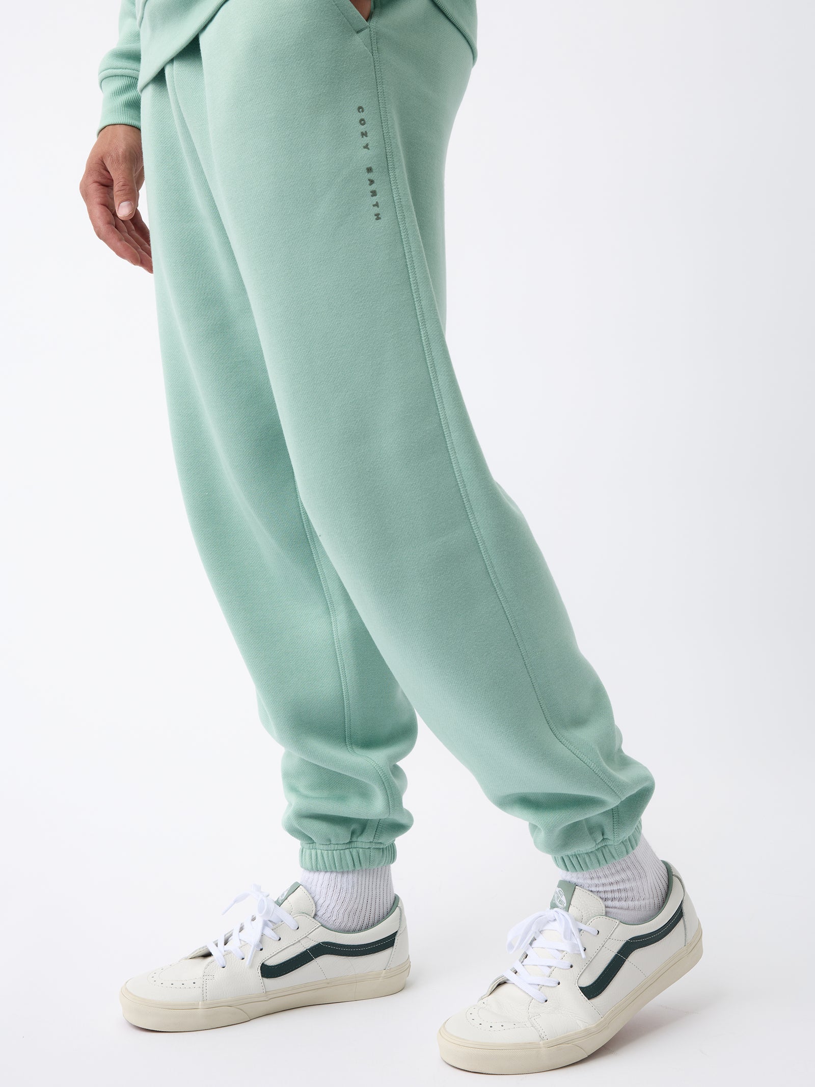 A person wearing Cozy Earth's Men's CityScape Sweatpant in pastel green, featuring subtle text detail on the side, stands casually. They complement the outfit with white sneakers accented in green and white socks against a plain white background. 