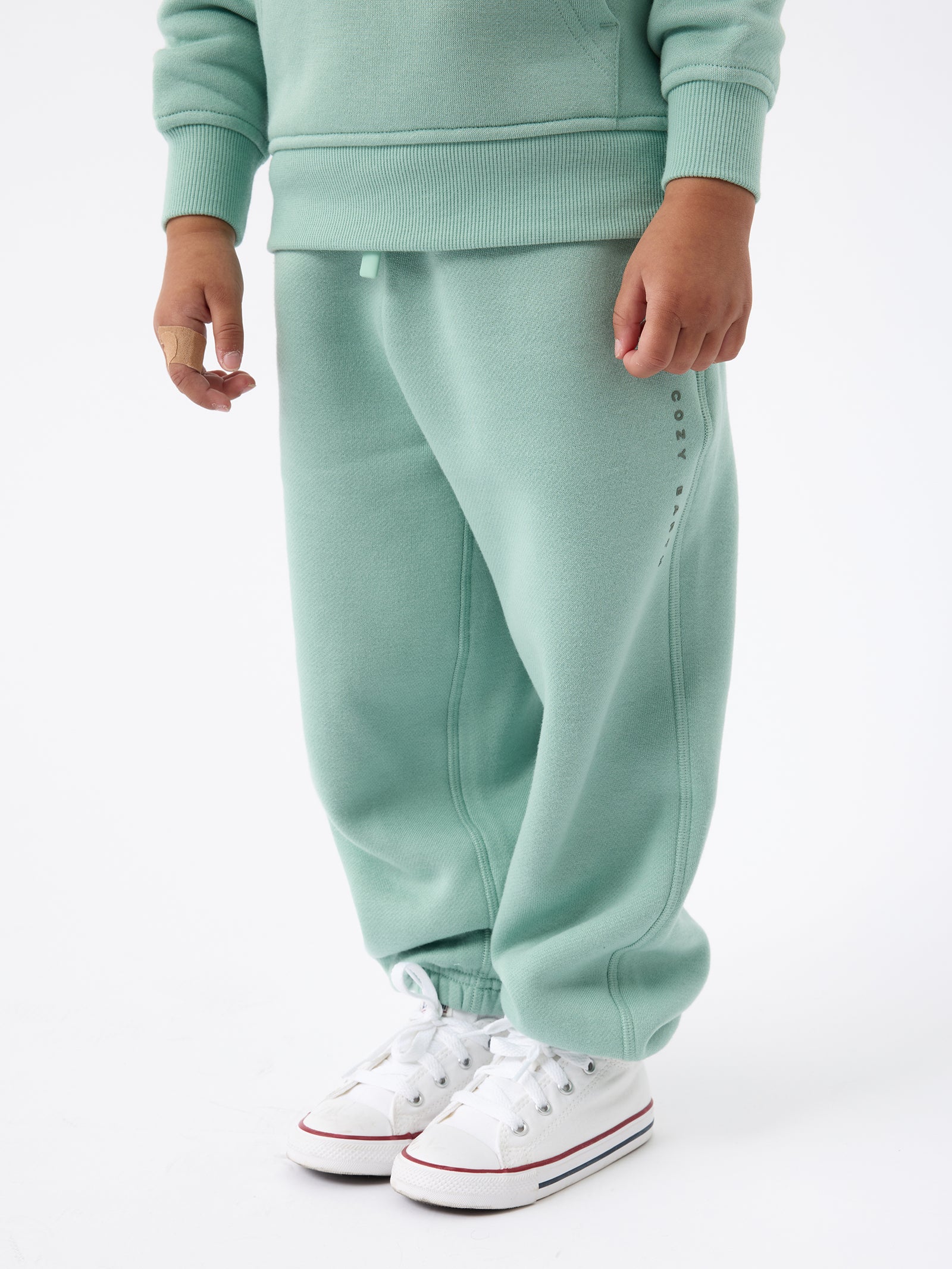 A child is wearing Cozy Earth's Kid's CityScape Sweatpant, a light green tracksuit that fits slightly oversized. They're paired with white sneakers and stand against a plain white background, with a bandage on their left index finger. The sweatpants feature subtle writing along the side. 