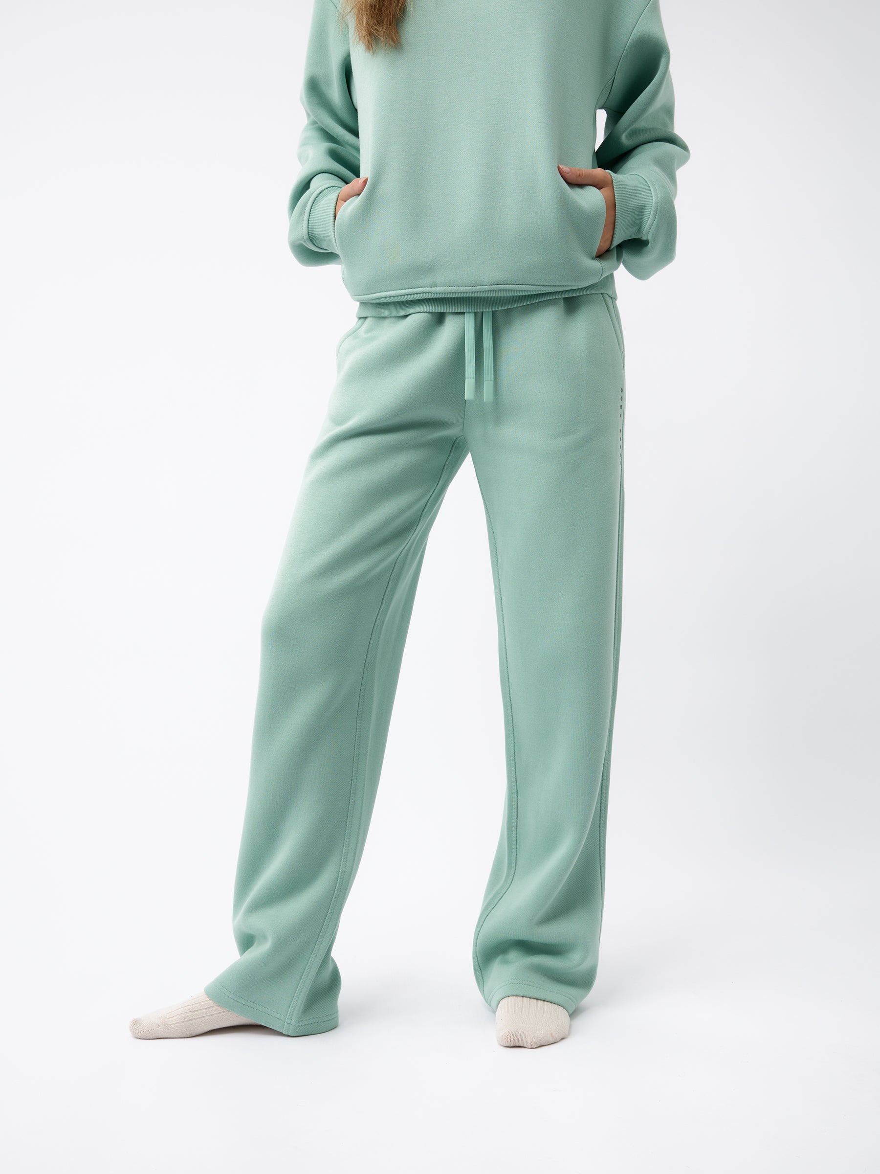 A person wearing the light green Women's CityScape Wide Leg Pant by Cozy Earth with hands in pockets. The outfit includes a long-sleeve top and matching wide-leg pants. The person is standing against a plain background, with beige socks visible. |Color:Wintergreen