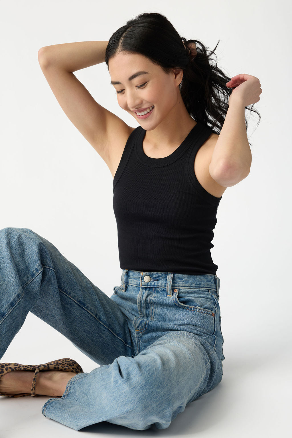 A dark haired woman seated on the floor wears Cozy Earth Women's Fine Ribbed Tank in Jet Black. She wears blue jeans and sits in front of a white background. 