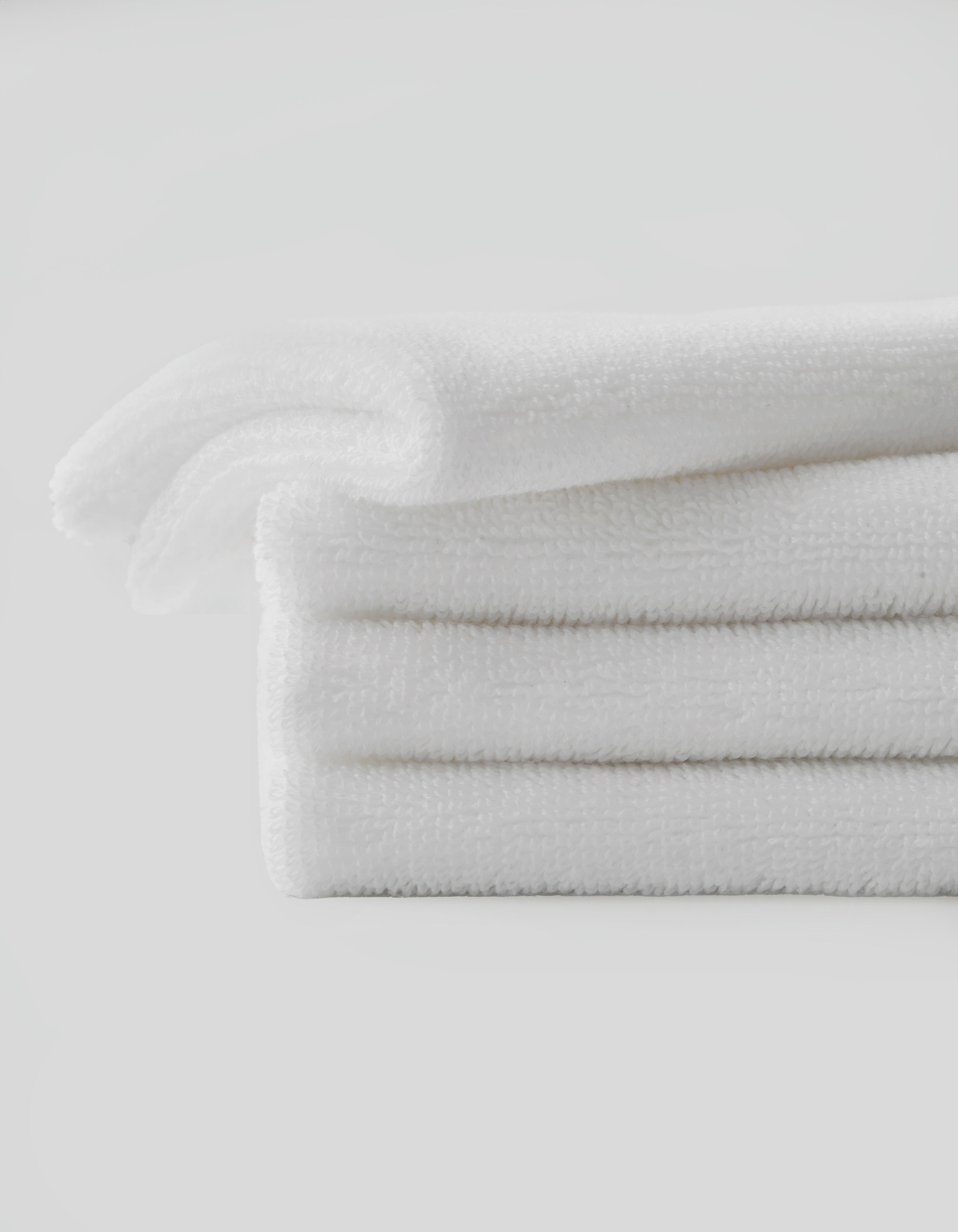 A neatly folded stack of three white towels from Cozy Earth's Revitalizing Pair set is placed against a plain white background. The towels appear soft and fluffy, with a clean, fresh look. The arrangement emphasizes their texture and pristine condition.