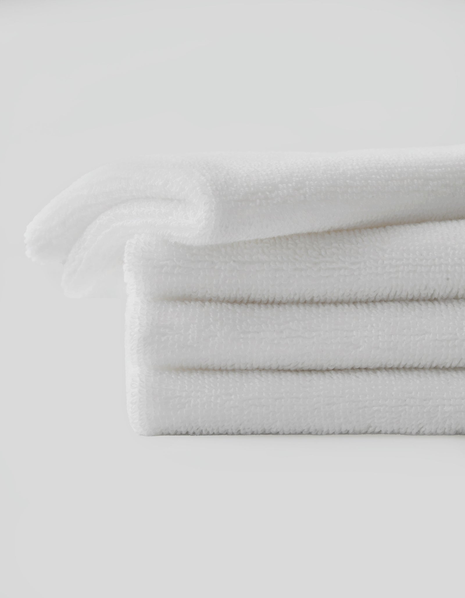 A stack of three neatly folded Complete Cleansing Cloths by Cozy Earth is placed against a plain white background. The white cloths appear soft and fluffy, suggesting a clean and fresh appearance. The overall image conveys a sense of cleanliness and simplicity.