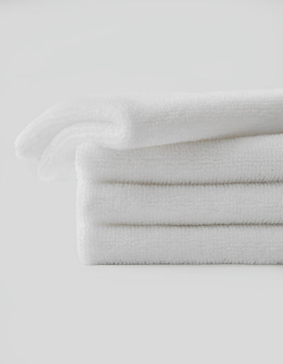 A stack of three neatly folded Complete Cleansing Cloths by Cozy Earth is placed against a plain white background. The white cloths appear soft and fluffy, suggesting a clean and fresh appearance. The overall image conveys a sense of cleanliness and simplicity.