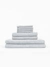 The Cozy Earth Nantucket Bath Towel Set features light grey textured towels in multiple sizes, neatly stacked from largest to smallest against a plain white background.|Color:Heathered Harbor Mist