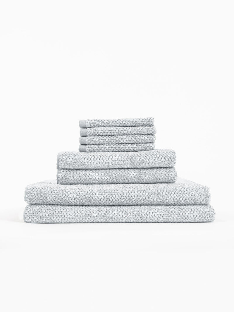 The Cozy Earth Nantucket Bath Towel Set features light grey textured towels in multiple sizes, neatly stacked from largest to smallest against a plain white background.