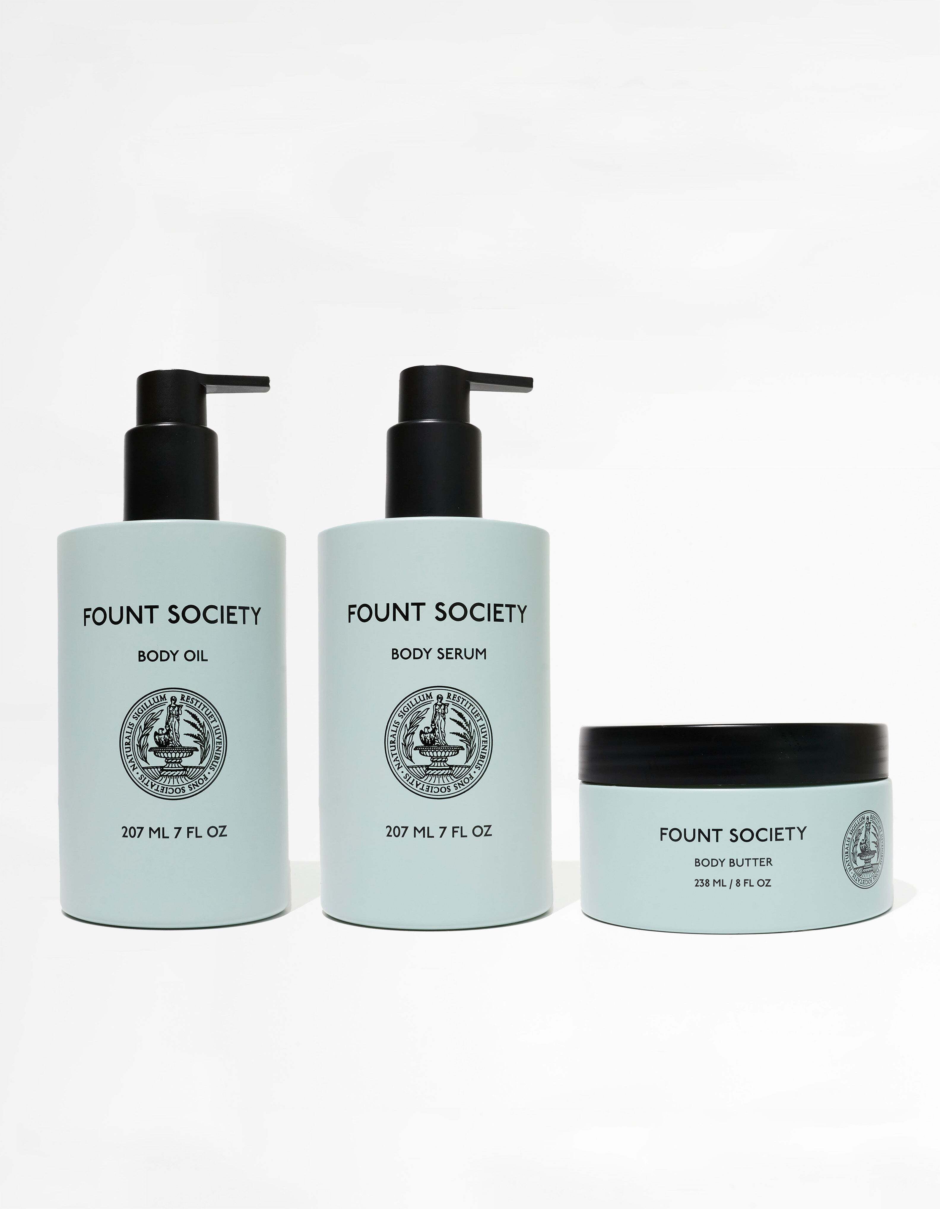 Image shows three skincare products from the brand Cozy Earth. From left to right: a bottle of Body Oil with a pump, a bottle of Body Serum with a pump, and a jar of Body Butter with a screw-on lid. All containers are light blue with black text and logos, collectively named the Moisture Trio.