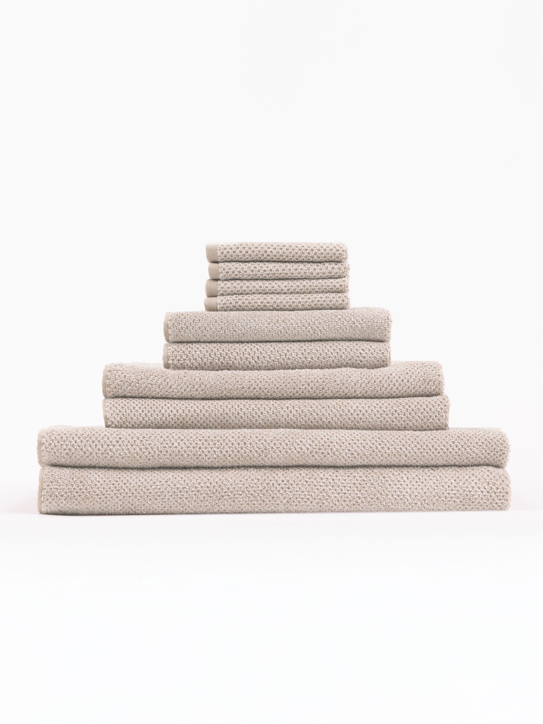 The Complete Nantucket Bath Bundle by Cozy Earth, featuring beige textured towels in various sizes, is neatly stacked pyramid-style on a white background.|Color:Heathered Sand