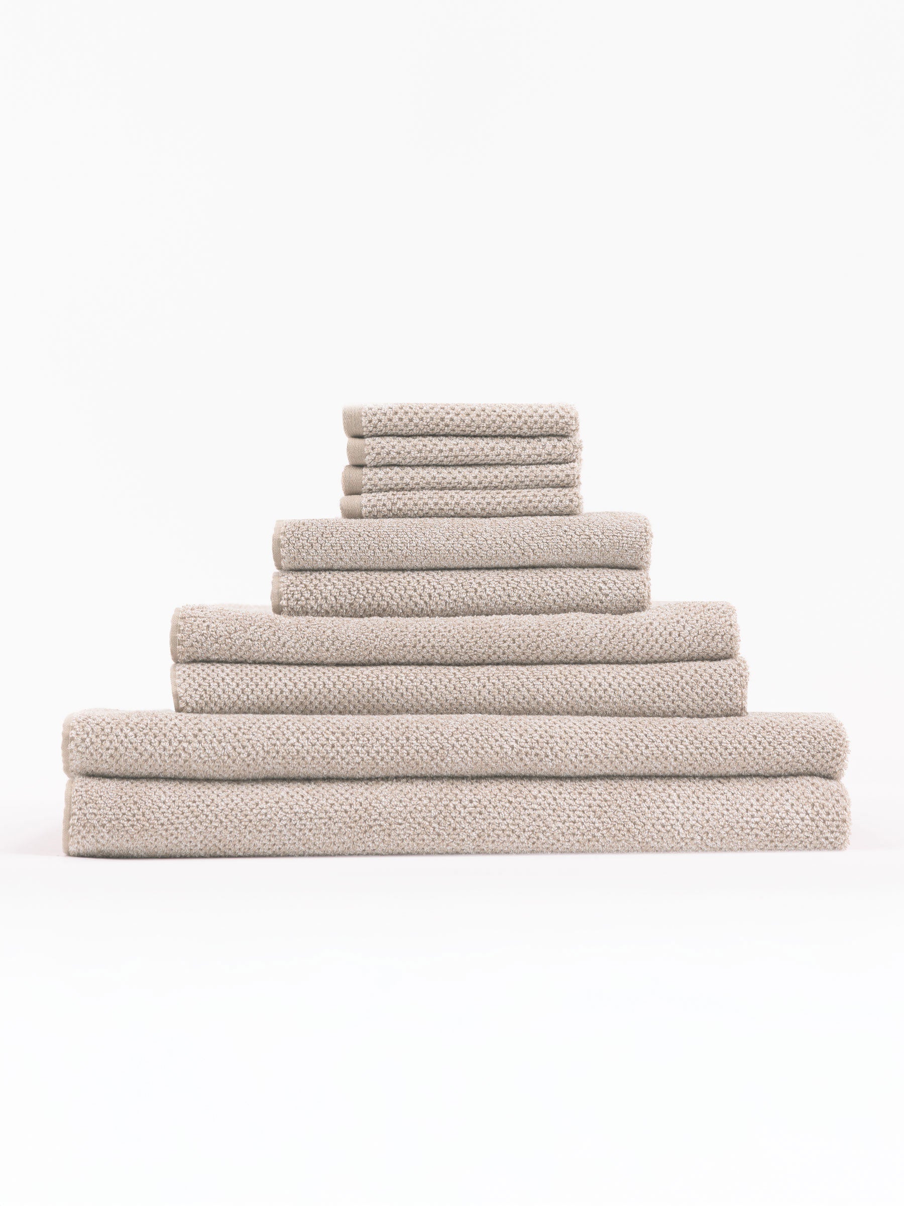 The Complete Nantucket Bath Bundle by Cozy Earth, featuring beige textured towels in various sizes, is neatly stacked pyramid-style on a white background.|Color:Heathered Sand