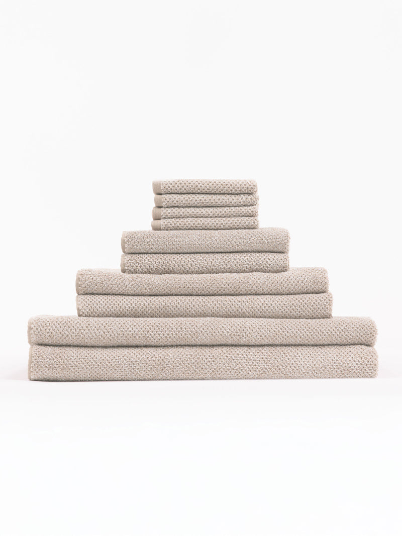 The Complete Nantucket Bath Bundle by Cozy Earth, featuring beige textured towels in various sizes, is neatly stacked pyramid-style on a white background.