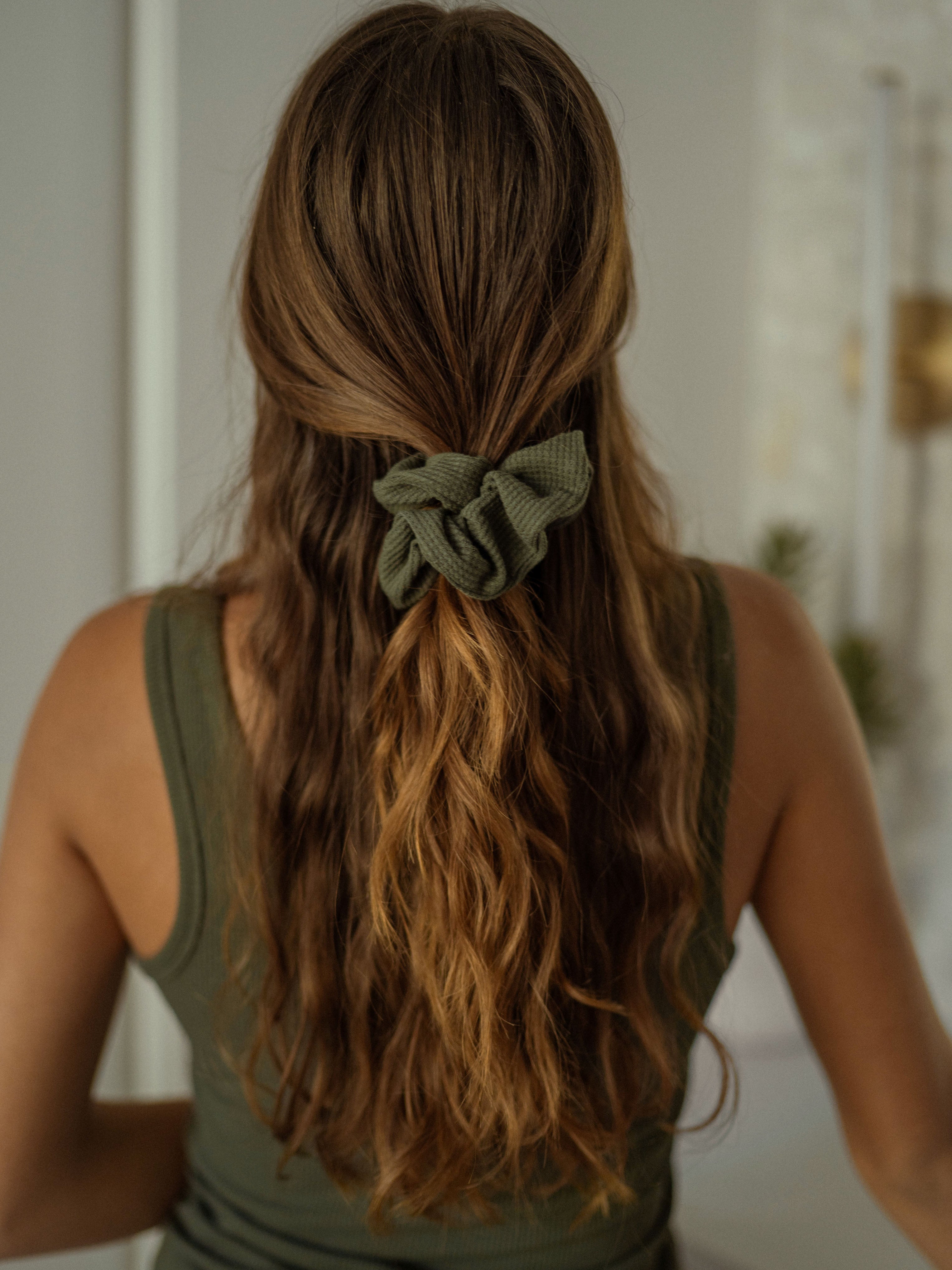 A woman with long, wavy brown hair is seen from the back, wearing a dark green tank top. Her hair is partially tied back with a Bamboo Waffle Knit Scrunchie from Cozy Earth, revealing a mix of light and dark tones. The background is blurred with neutral and light colors.