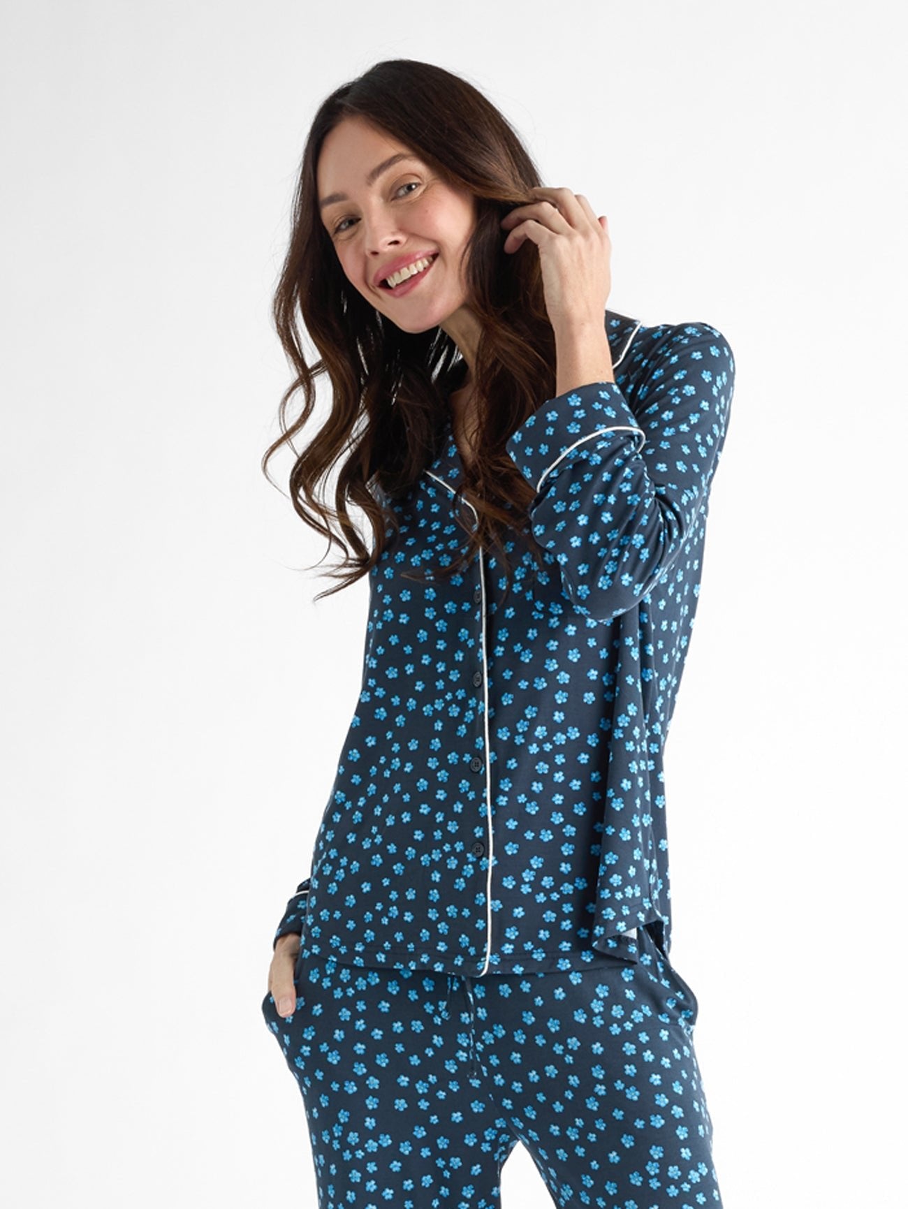 A smiling person in Cozy Earth Women's Bamboo Stretch-Knit Long Sleeve Pajama Top, featuring a dark blue fabric with small blue floral patterns, stands with one hand in their pocket against a white backdrop. 