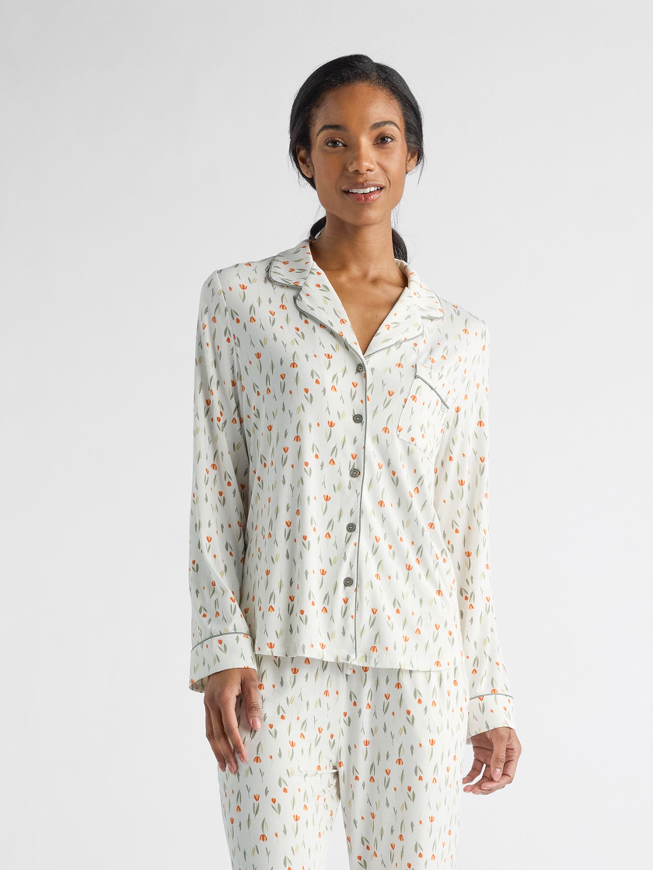 A person is wearing a Women's Bamboo Stretch-Knit Long Sleeve Pajama Top by Cozy Earth, which features a light base color and small red and orange shapes, complete with a collar and buttons. The background is plain white. 