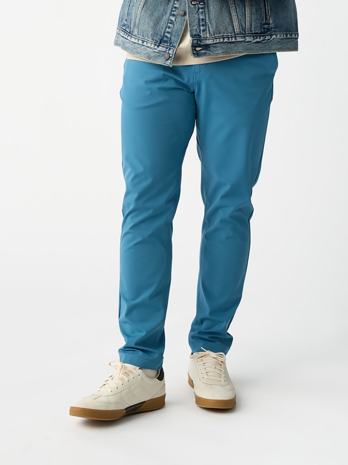 A person stands against a plain white background, wearing the Men's Everywhere Pant in blue from Cozy Earth, paired with beige sneakers and a denim jacket. The focus is on the lower half of the body. |Color:Wave