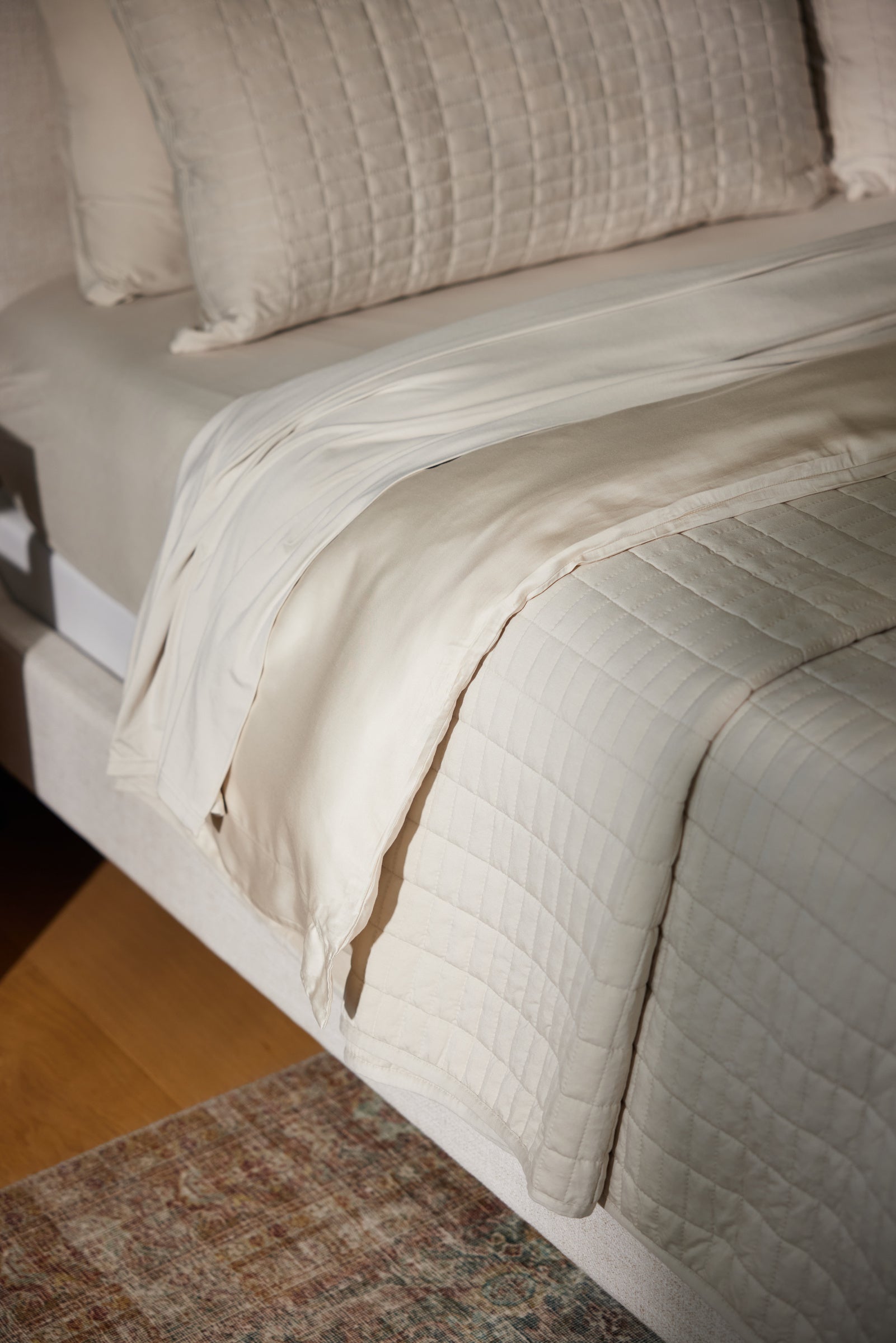 A neatly made bed features Cozy Earth's Bamboo Jersey Fitted Sheet in cream, with a quilted blanket and matching pillows atop a beige upholstered frame, next to a patterned rug on a wooden floor.|Color:Birch