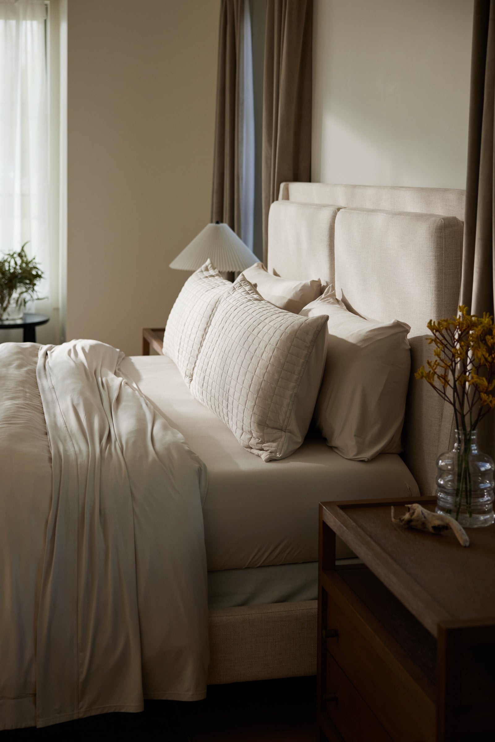 A bed with beige Bamboo Jersey Fitted Sheet by Cozy Earth and large pillows sits beside a wooden nightstand with a lamp and a vase of yellow flowers, illuminated by soft natural light from the window.|Color:Birch