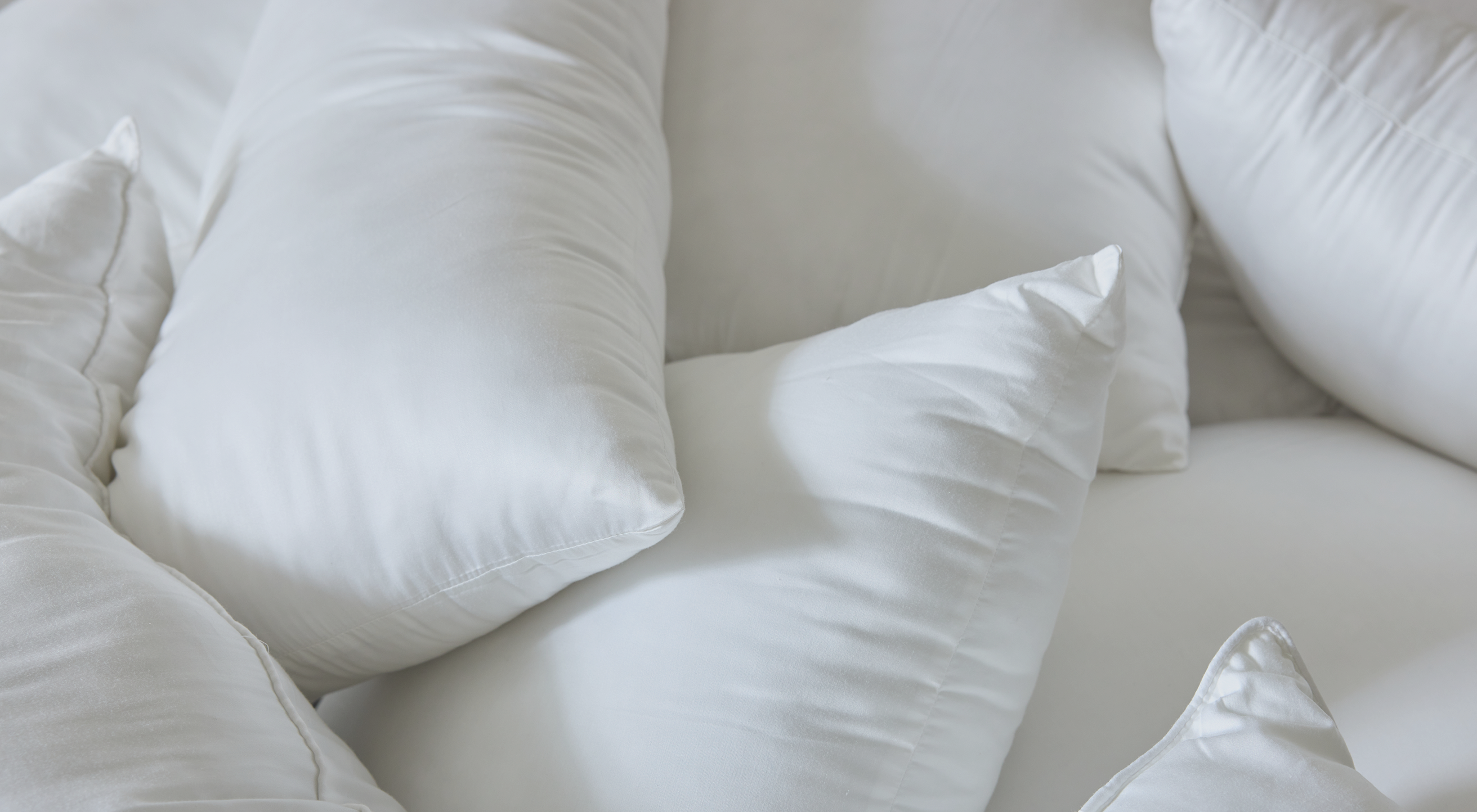 A close-up view of several fluffy white pillows stacked together. The pillows have a soft, smooth texture, with plump and inviting shapes, creating a cozy and comfortable appearance.