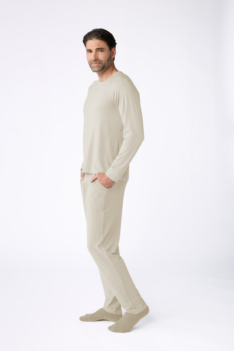 A man wears the Men's Loews Hotel Lounge Set by Cozy Earth—matching light beige long-sleeve top, pants, and socks—posing in a relaxed manner against a clean white background for a minimal look.