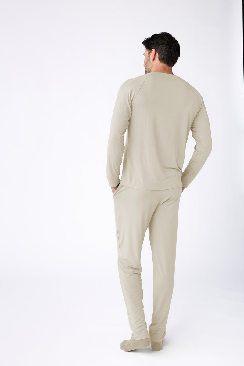 A man with dark hair faces away from the camera, wearing Cozy Earth's Men's Loews Hotel Lounge Set in light beige with matching long-sleeve top, pants, and beige socks, set against a plain white background.