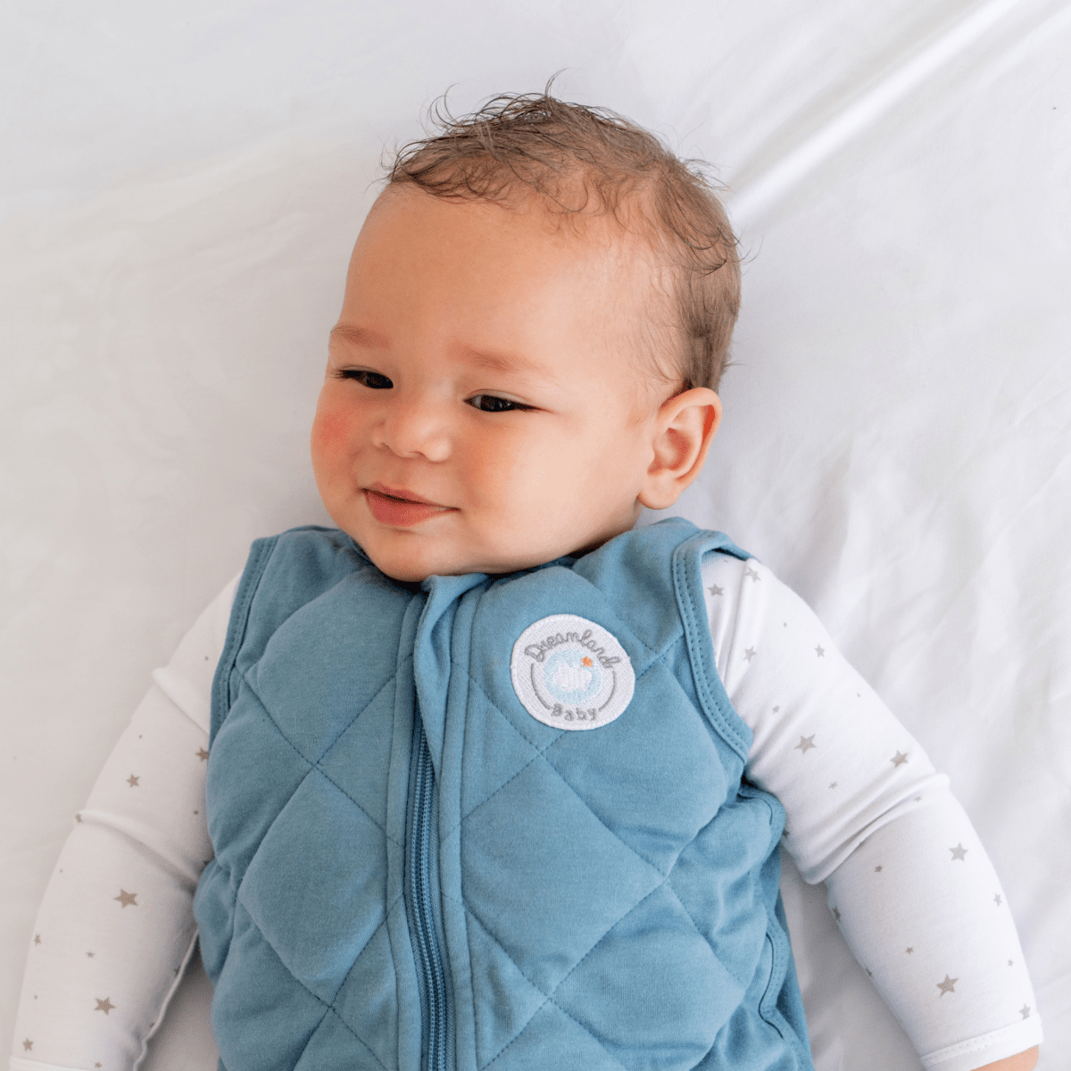 A smiling baby with curly hair wears the Dreamland Baby Dream Weighted Sleep Sack and a white onesie with gray stars, lying on a white blanket.
