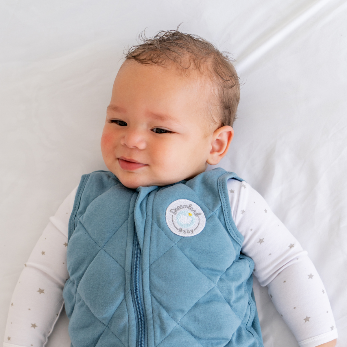 A smiling baby with curly hair wears the Dreamland Baby Dream Weighted Sleep Sack and a white onesie with gray stars, lying on a white blanket.