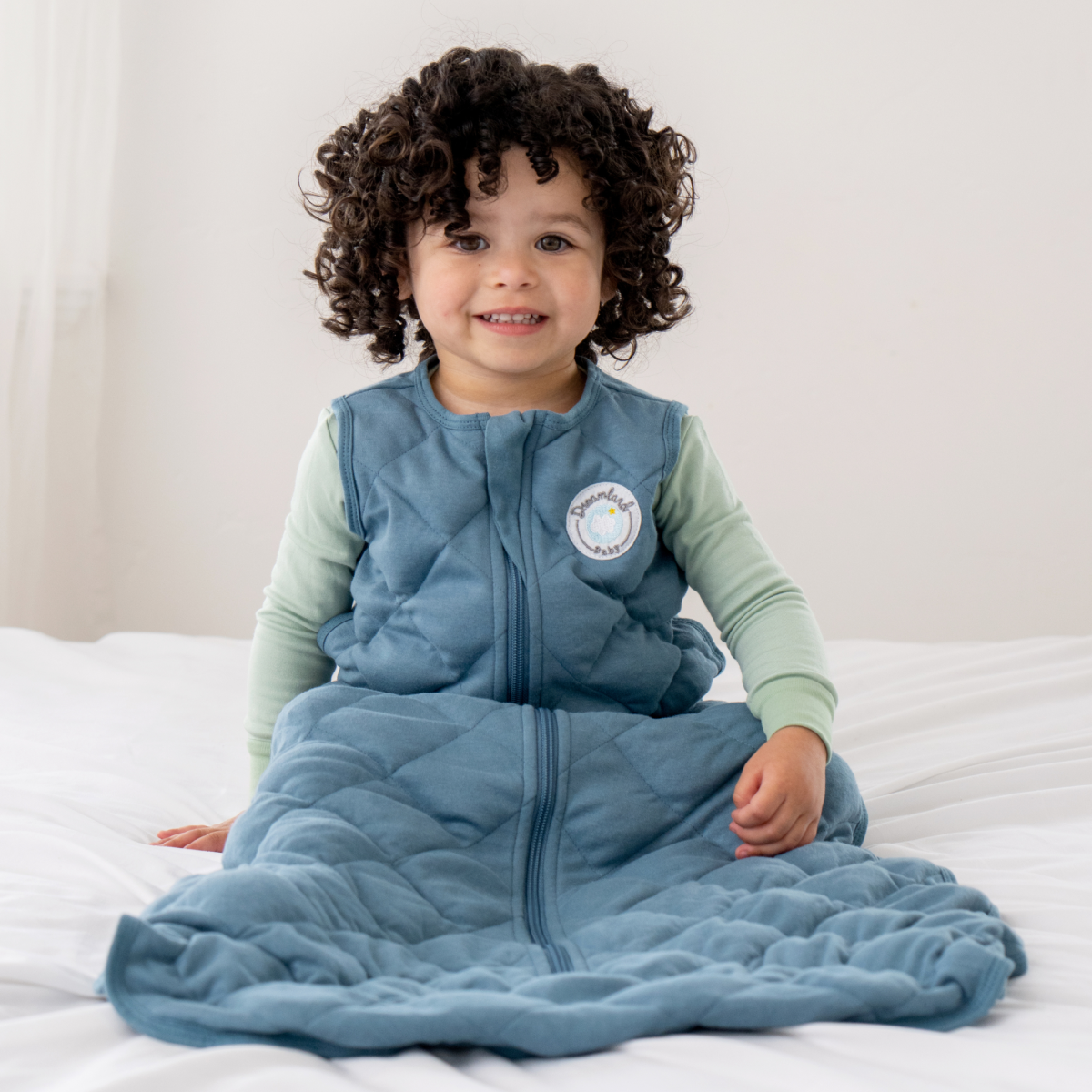A young child with curly hair smiles on a bed, wearing the Dreamland Baby Dream Weighted Sleep Sack in blue over light green pajamas. The sack features a circular logo on the chest.