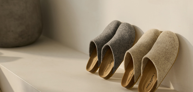 Two pairs of woolen slippers in gray and beige are neatly lined up on a white surface. The light casts soft shadows, creating a cozy and minimalist atmosphere.