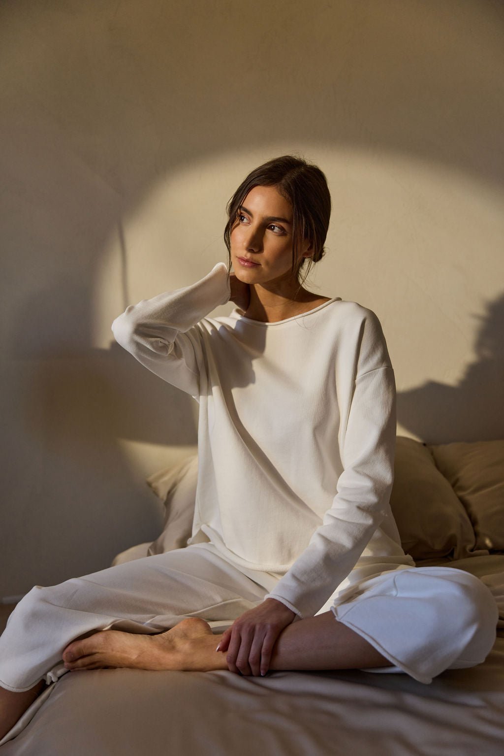 A person sits cross-legged on a bed wearing Cozy Earth's Women's Chelsea Pullover, gazing thoughtfully to the side with one hand on their neck. Warm sunlight casts gentle shadows on the wall behind them. 