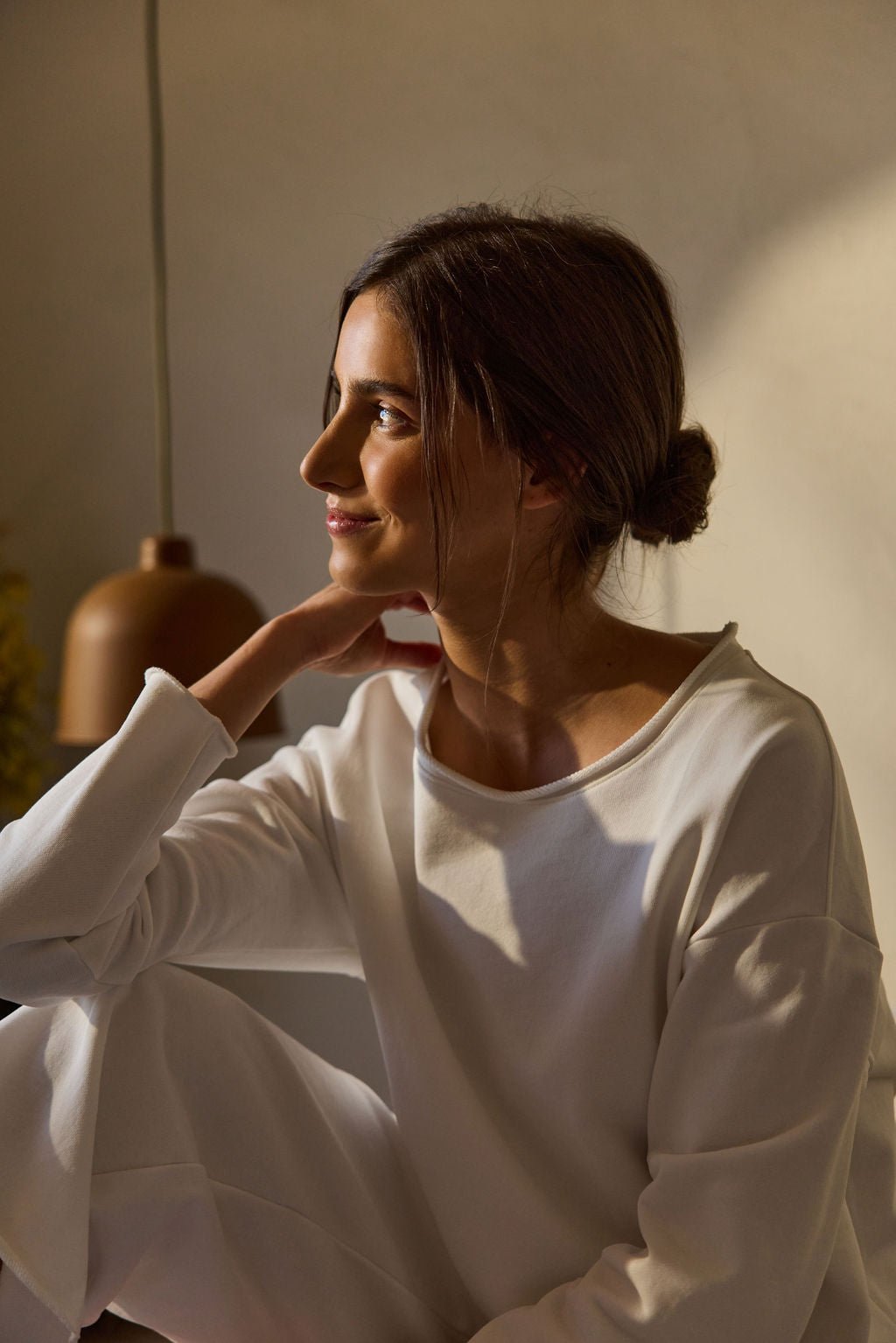 In soft, warm lighting, a woman with long brown hair in a bun sits wearing the Cozy Earth Women's Chelsea Pullover and matching pants. She gently smiles while looking to her left. A brown pendant light is visible in the background. 