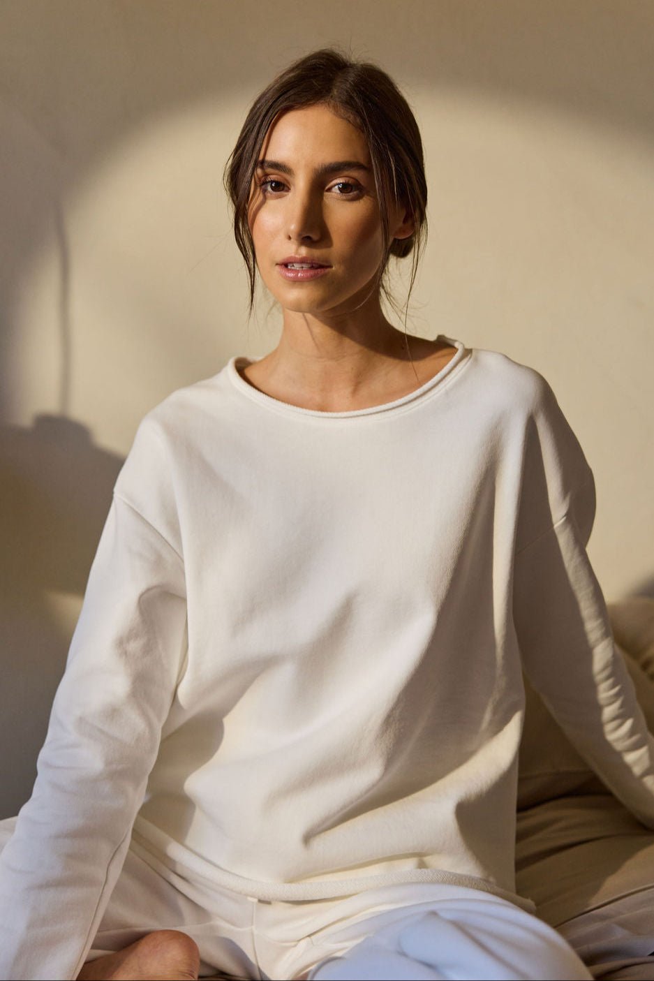 A person with long brown hair sits on a bed, wearing the Women's Chelsea Pullover by Cozy Earth and light-colored pants. The background has a warm, soft focus, creating a relaxed and serene atmosphere. |Color:Bone