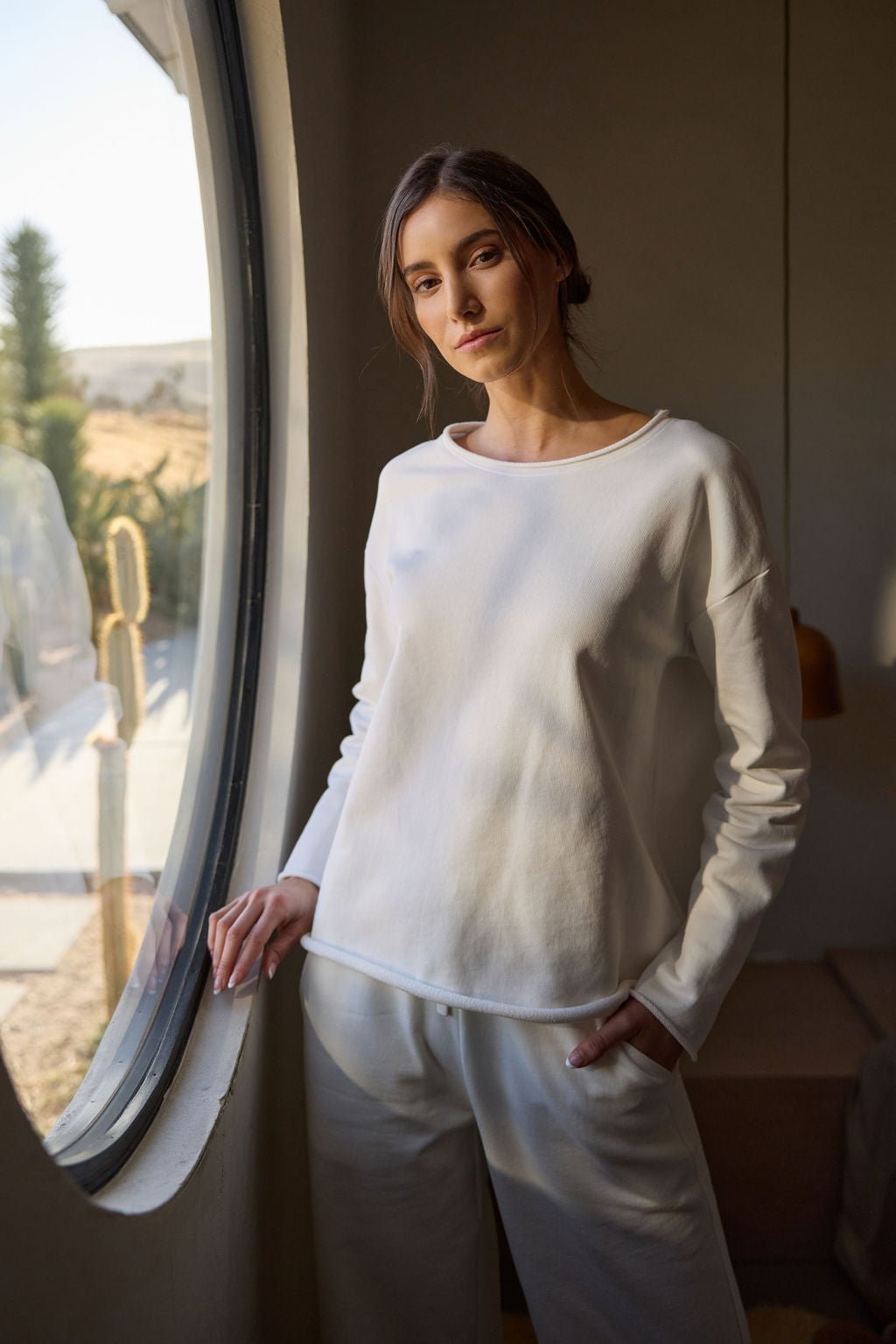 A person stands by a large round window, wearing the Cozy Earth Women's Chelsea Pullover and casual pants. They look calmly at the camera. The room is warmly lit, with a desert landscape visible outside. 