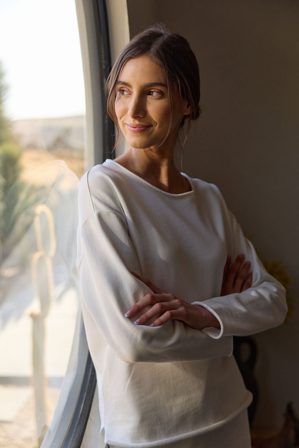 Wearing a Women's Chelsea Pullover from Cozy Earth, a woman stands by a window with her arms crossed, smiling and looking to the side as sunlight streams in, illuminating her face and creating a warm atmosphere. 