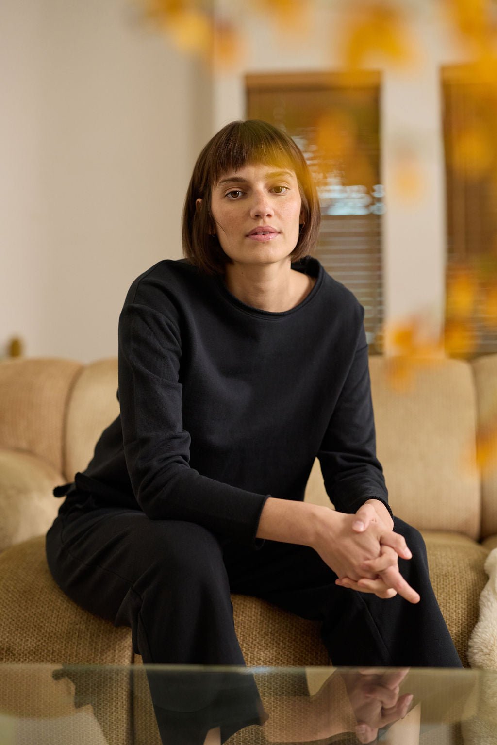 A person with short brown hair sits on a beige sofa, leaning forward with hands clasped. They wear the Women's Chelsea Pullover by Cozy Earth in dark tones, paired with matching pants. The room features neutral colors and a window in the background. 