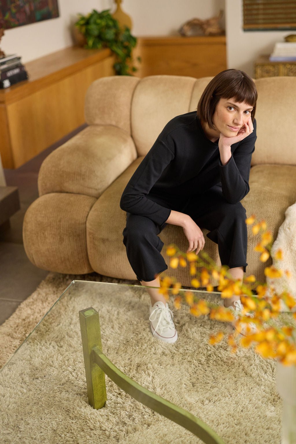 A short-haired person sits on a beige sofa, leaning forward with their chin on their hand. They wear a black outfit and white sneakers, identified as the Women's Chelsea Pullover by Cozy Earth. In the foreground is a glass coffee table adorned with vibrant yellow flowers.  