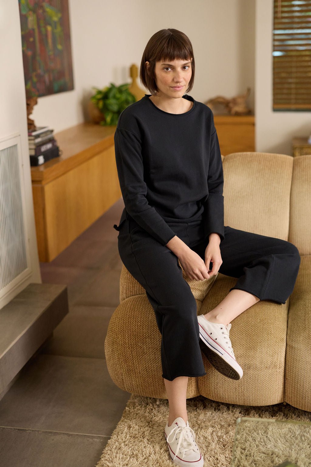 A person with short hair sits on an armrest in a cozy living room, wearing a black Women's Chelsea Pullover by Cozy Earth and white sneakers. The background features a painting, books, plants on a wooden sideboard, earthy tones, and a rug.  