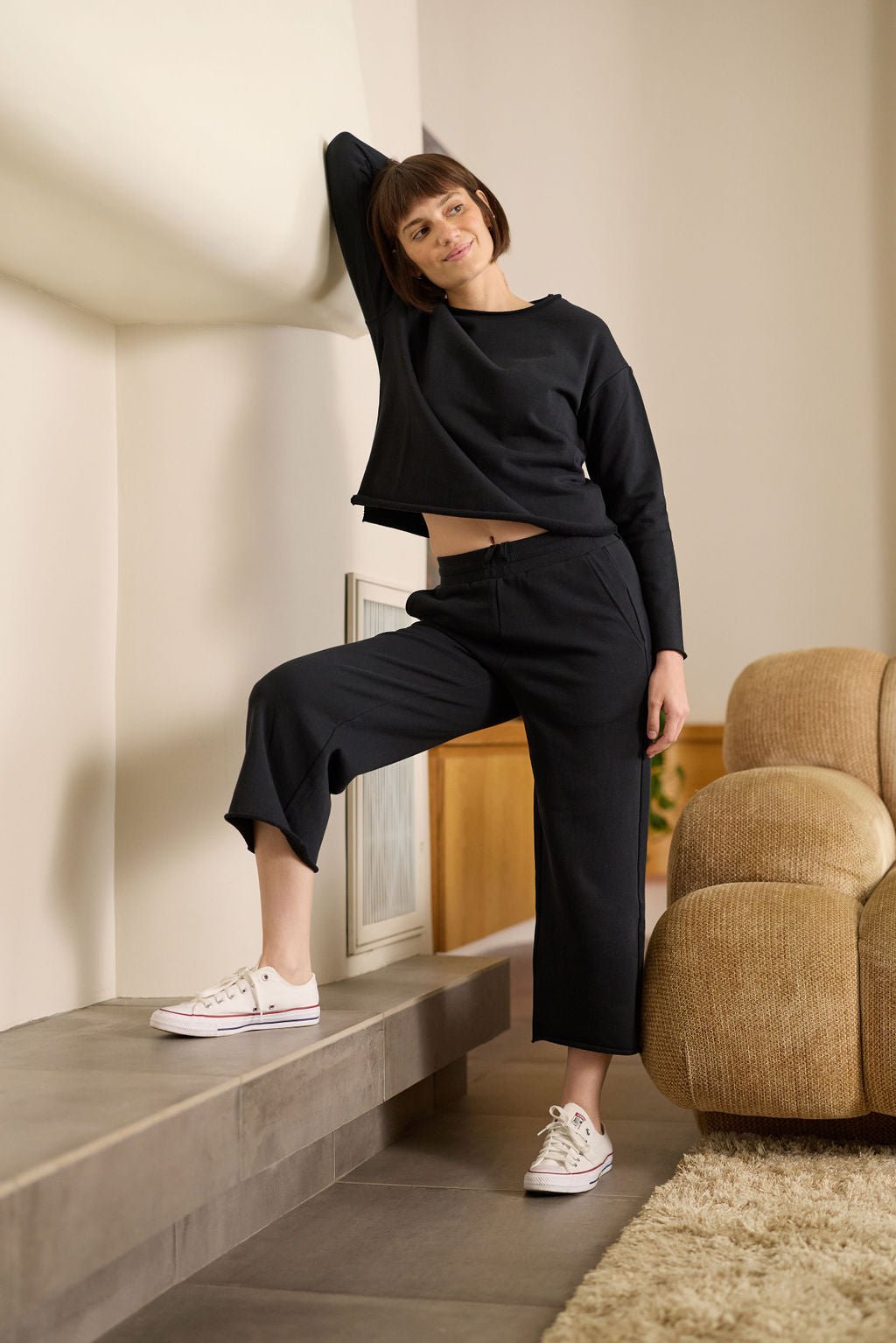 A person with short brown hair leans against a wall indoors, smiling and posing in Cozy Earth's Women's Chelsea Pullover, paired with matching wide-legged pants and white sneakers. One leg is bent and resting on a platform. 