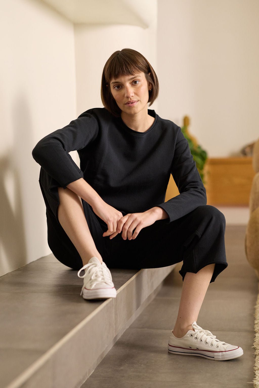 A person with short brown hair sits casually on a ledge, dressed in Cozy Earth's Women's Chelsea Pullover and black pants, wearing white sneakers. The softly lit interior background has hints of greenery and wood. 