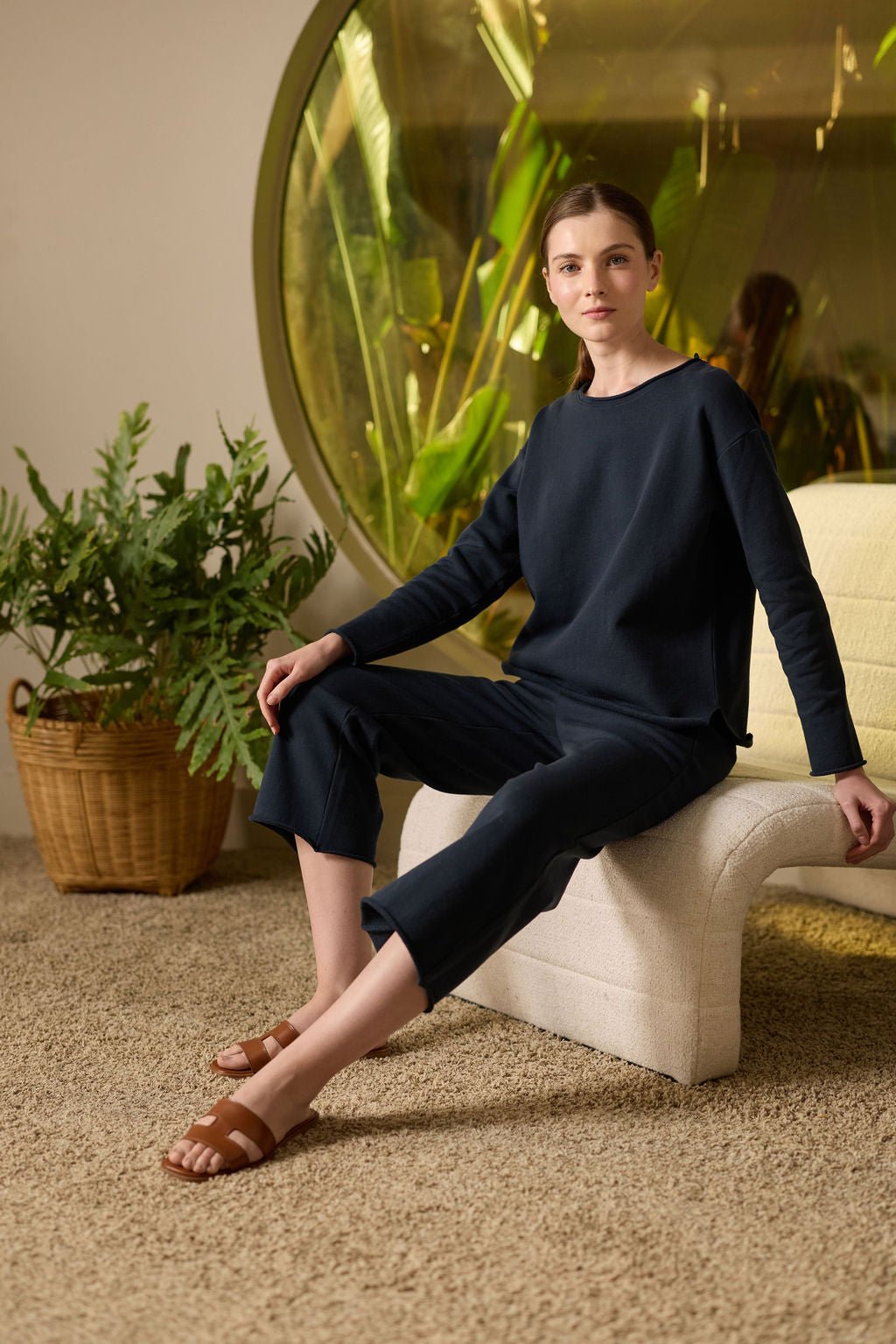 A person with long hair sits on a cream chair, wearing Cozy Earth's Women's Chelsea Pullover and pants. They are in a room with a large circular window showing greenery outside, next to a potted plant on the carpeted floor. 