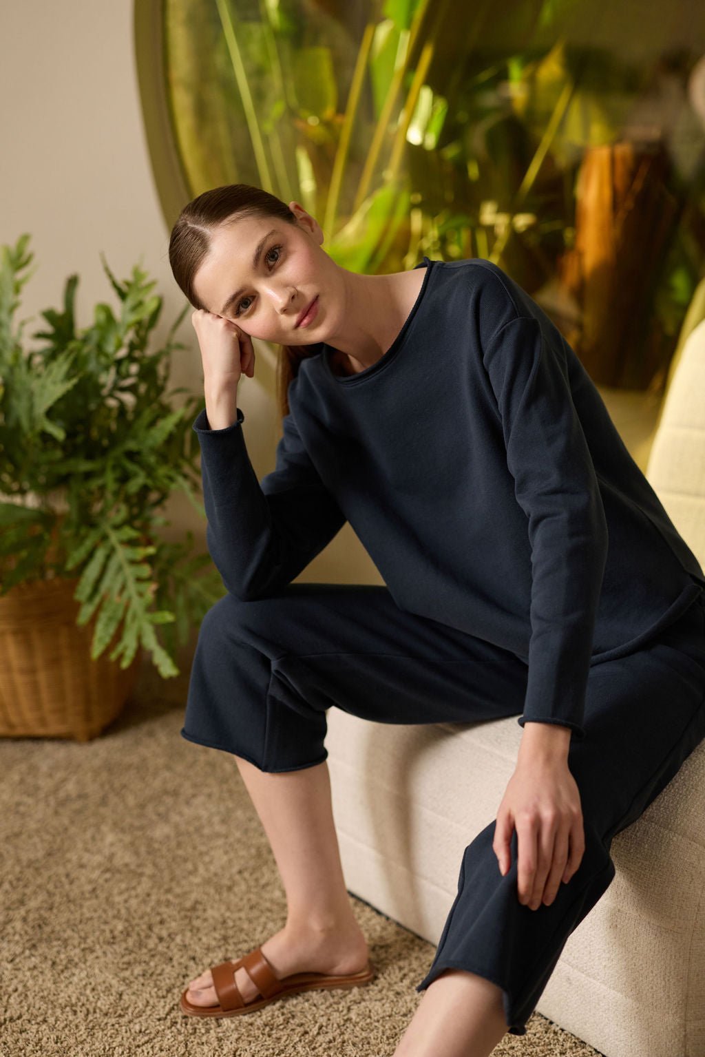 A person with brown hair, clad in a Cozy Earth's Women's Chelsea Pullover and brown sandals, sits indoors on a white sofa with one elbow on their knee. Green foliage and a potted plant can be seen in the background. 