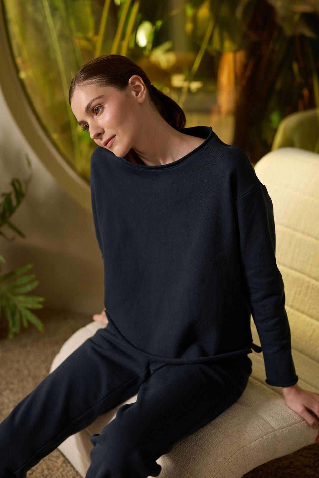 A woman with long brown hair is seated on a cream-colored chair, wearing a Cozy Earth Women's Chelsea Pullover in dark blue with matching pants. The indoor setting has large windows and green plants in the background. 