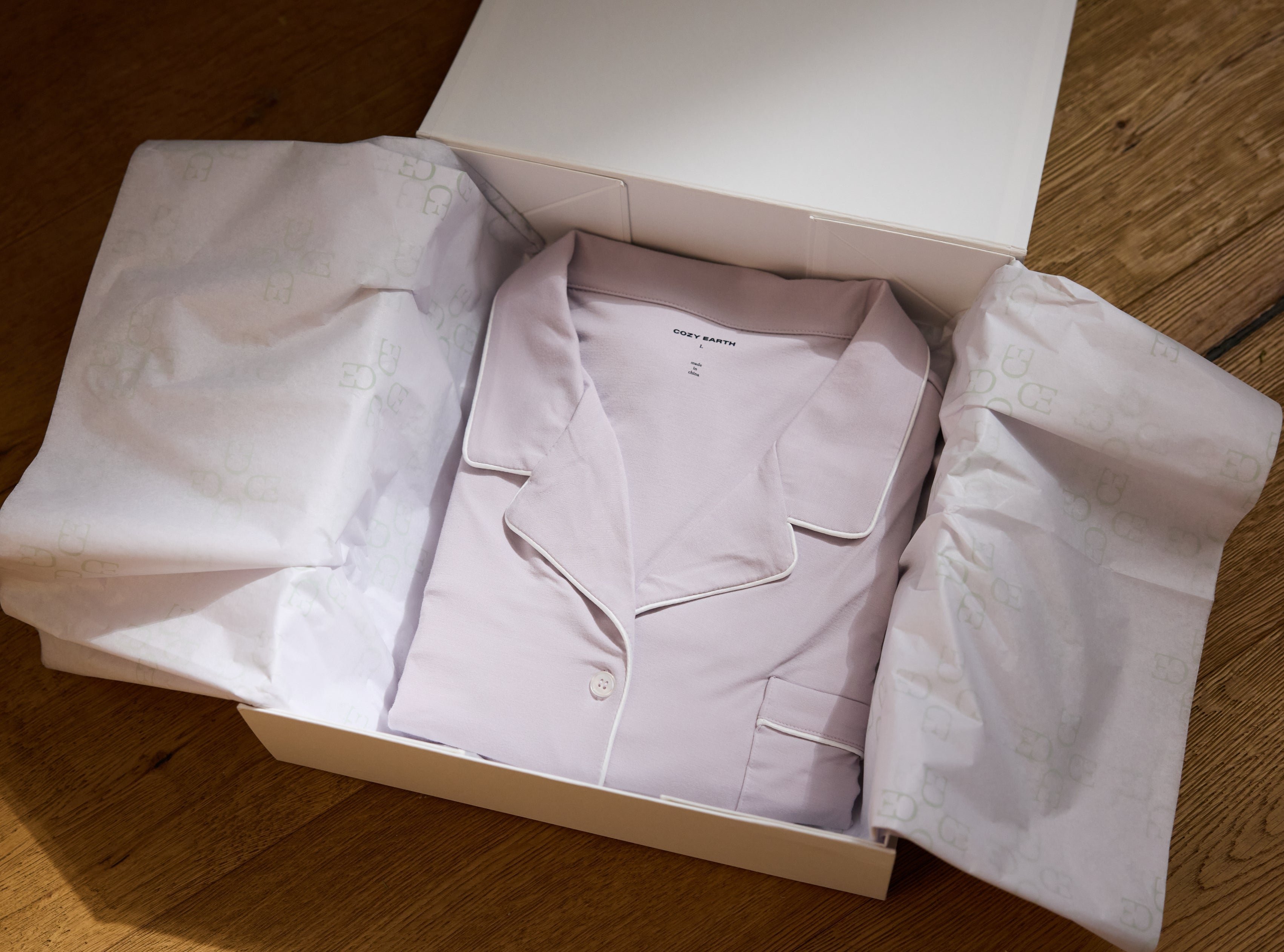 A neatly folded light pink pajama set with white piping is displayed inside an open white gift box lined with printed tissue paper. The box is placed on a wooden floor.