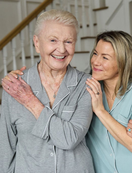 An elderly woman and a younger woman smile warmly. They are wearing pajamas; the elderly woman in grey and the younger in blue. They stand closely in a cozy home setting with a staircase in the background.