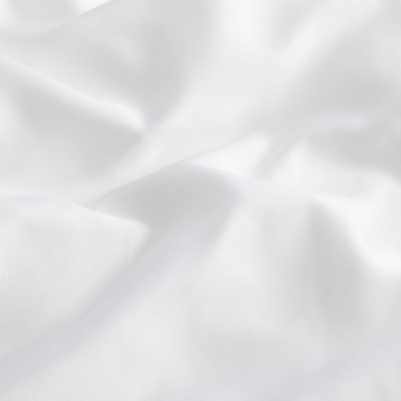 The Silk Comforter by Cozy Earth is crafted from a soft, smooth white fabric that gently folds and creases, offering a serene and clean appearance. Its evenly lit surface accentuates the texture and contours of the material.