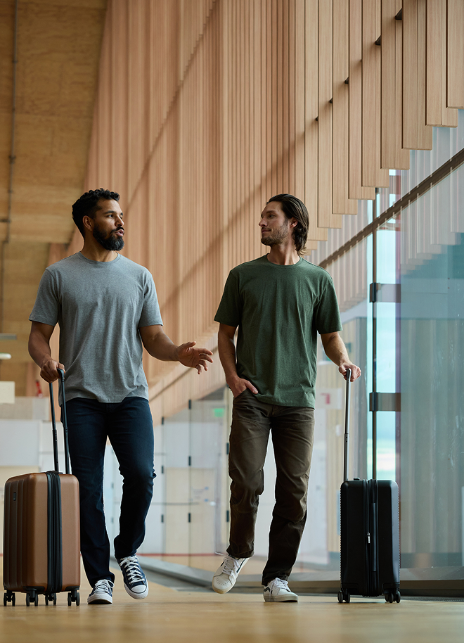 Two men walk side by side in an airport terminal. Each is pulling a suitcase. One wears a gray shirt and jeans, the other a green shirt and brown pants. They appear to be engaged in conversation, with wooden panels lining the corridor.